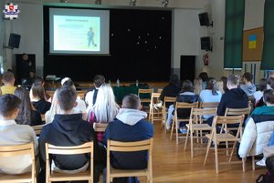 na zdjęciu dzielnicowy który prowadzi zajęcia z młodzieżą i osoby siedzące na krzesłach a przed nimi rzutnik z prezentacja