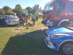 zdjęcie przedstawia radiowóz policyjny po prawej i wóz strażacki a po lewej stronie widać strażaków udzielających pomocy poszkodowanym w wypadku drogowym