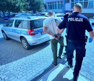na zdjęciu zatrzymany mężczyzna razem z doprowadzającymi go policjantami przy radiowozie