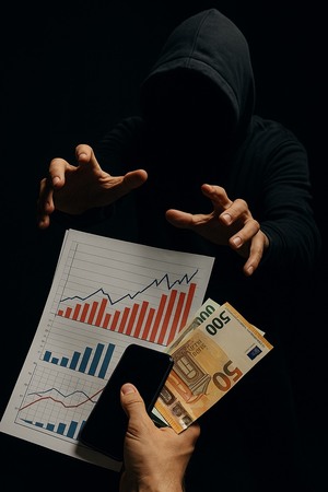 oszust w ciemnym kapturze przed nim dłonie, wykres i banknoty