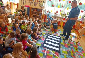 na zdjęciu dzielnicowy,który stoi przed dziećmi, dzieci po lewej siedzą na dywanie